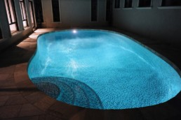 Freeform indoor swimming pool photographed at night time. The water glows from the pool light and showcases the blue mosaic glass tile.